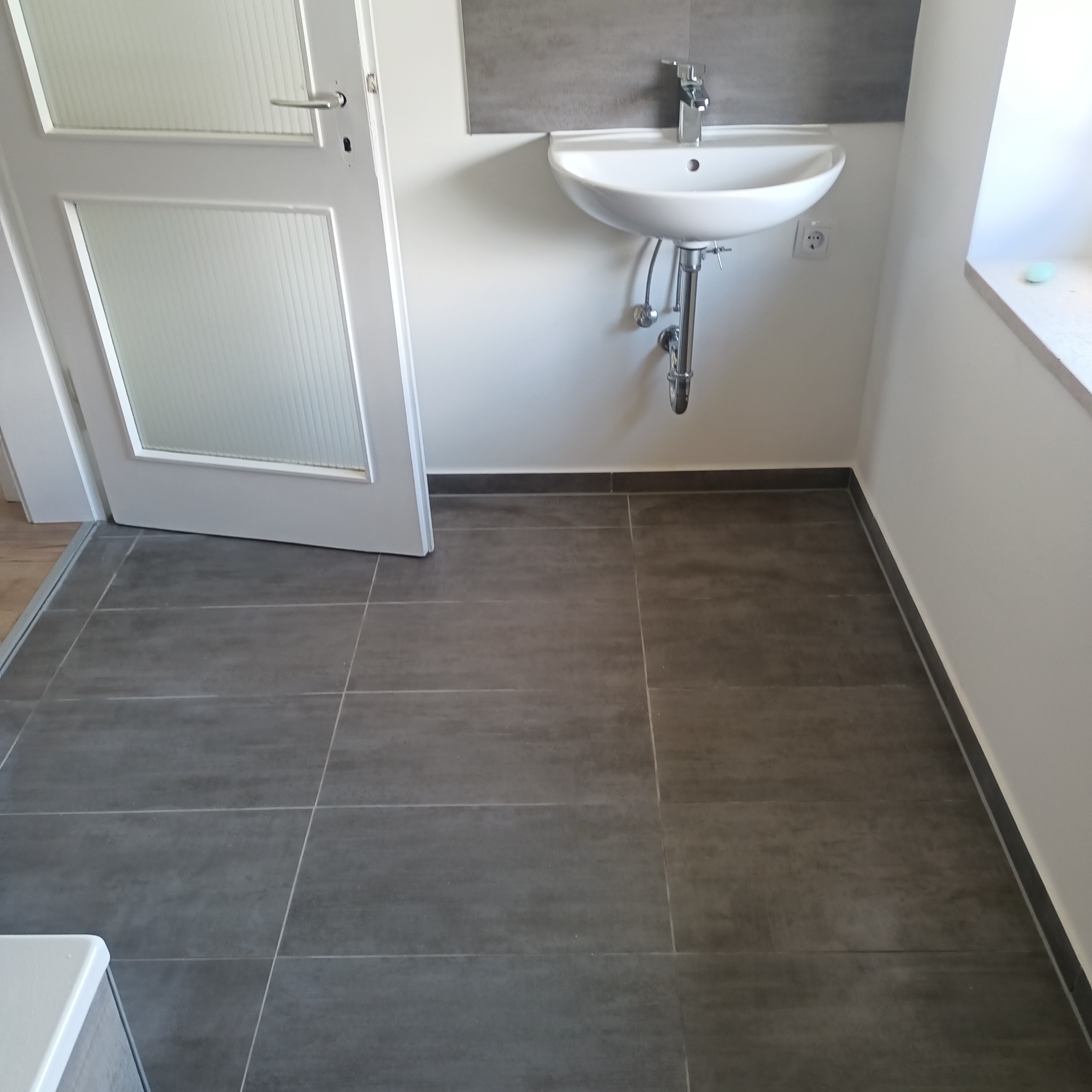 Nowoczesna łazienka z szarą podłogą z płytek, białą umywalką i drzwiami z przeszkleniami. Minimalistyczny design z akcentem na proste linie i funkcjonalność.