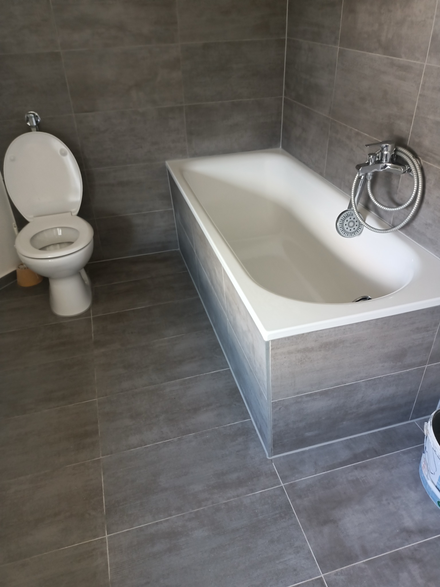 Nowoczesna łazienka z szarymi płytkami: biała wanna z chromowaną armaturą i toaleta. Minimalistyczny design, świeżo po remoncie. Widoczny fragment wiadra.