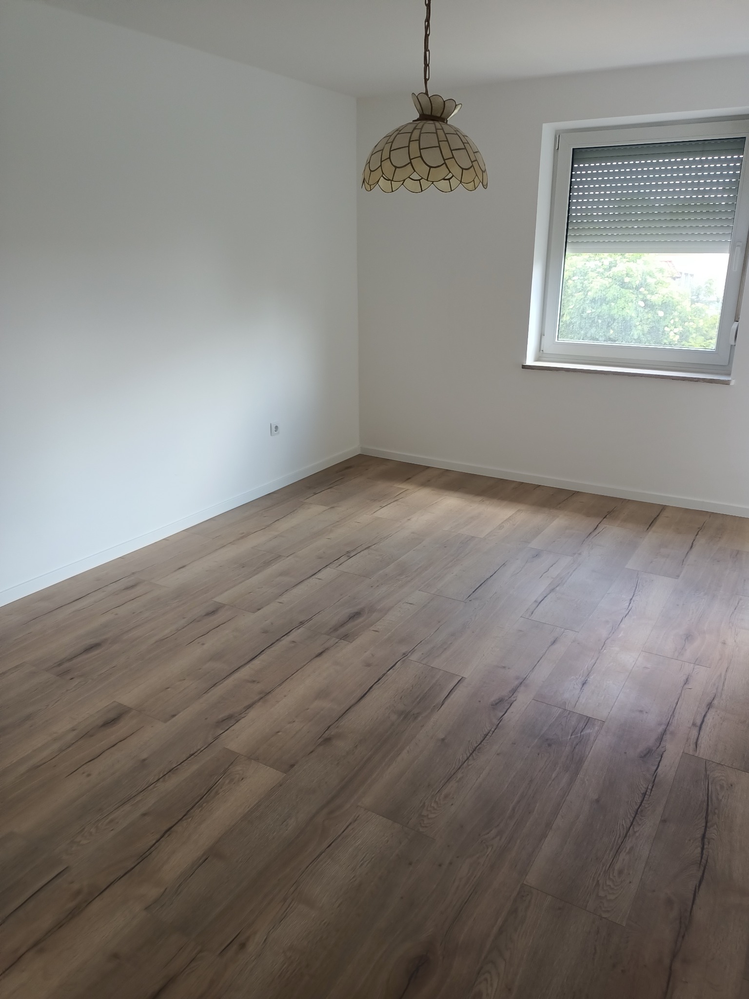 Jasne wnętrze pokoju po remoncie: panele podłogowe imitujące drewno, białe ściany, okno z roletami i lampa w stylu retro. Widok z perspektywy podłogi.