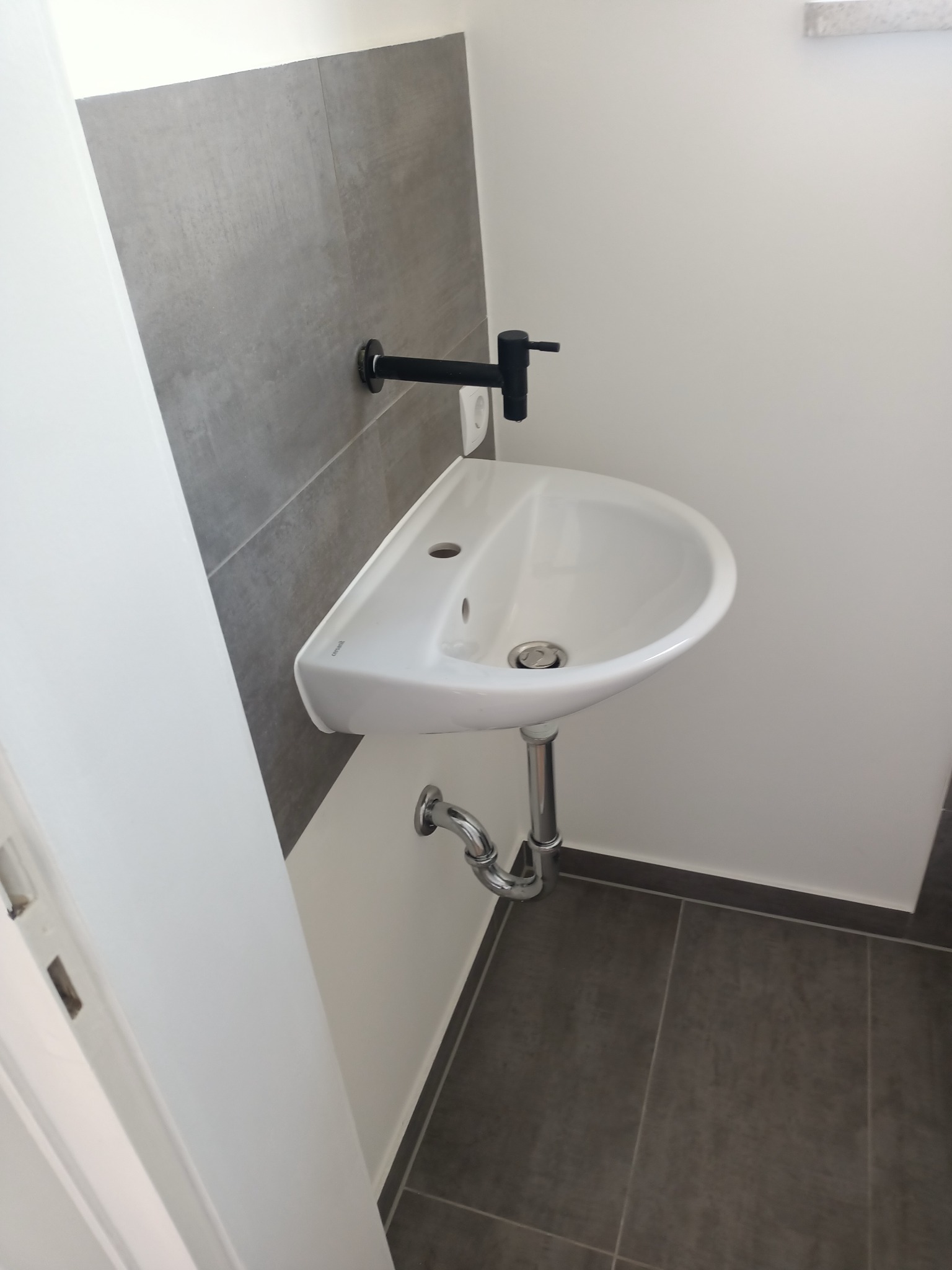 Biała umywalka z czarną armaturą na tle szarych płytek w łazience po remoncie w Rawiczu. Widoczny syfon i fragment podłogi z ciemnych płytek.