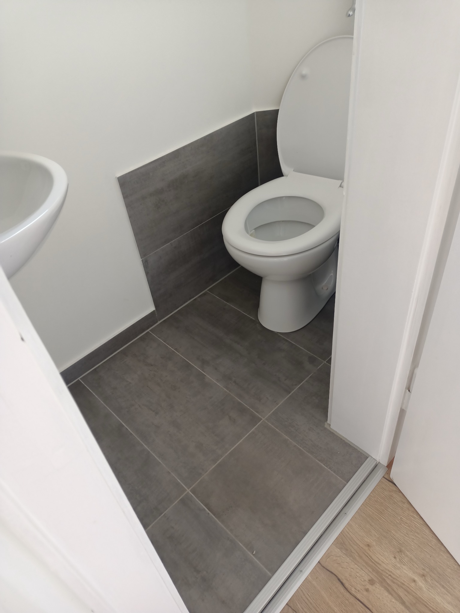 Jasna łazienka po remoncie: szare płytki podłogowe i ścienne, biała toaleta z otwartą klapą, umywalka i białe ściany. Widok zza uchylonych drzwi.