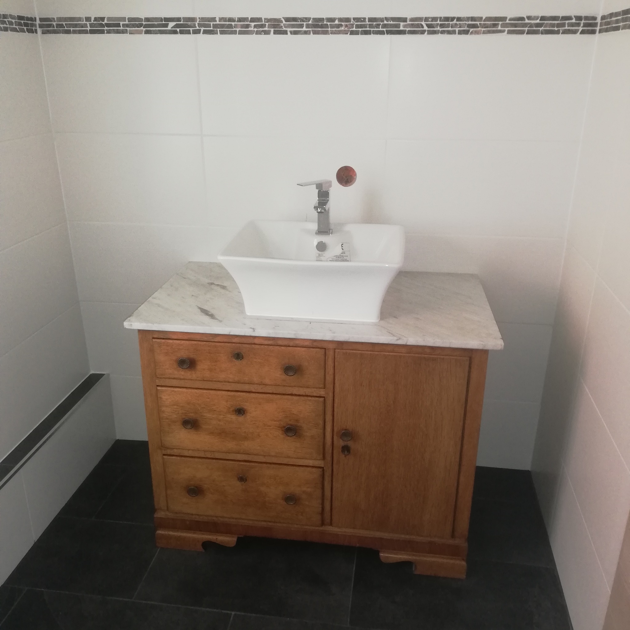 Wykończona łazienka z drewnianą szafką pod umywalką i białą, kwadratową umywalką. Na ścianach białe płytki z dekoracyjnym pasem mozaiki.