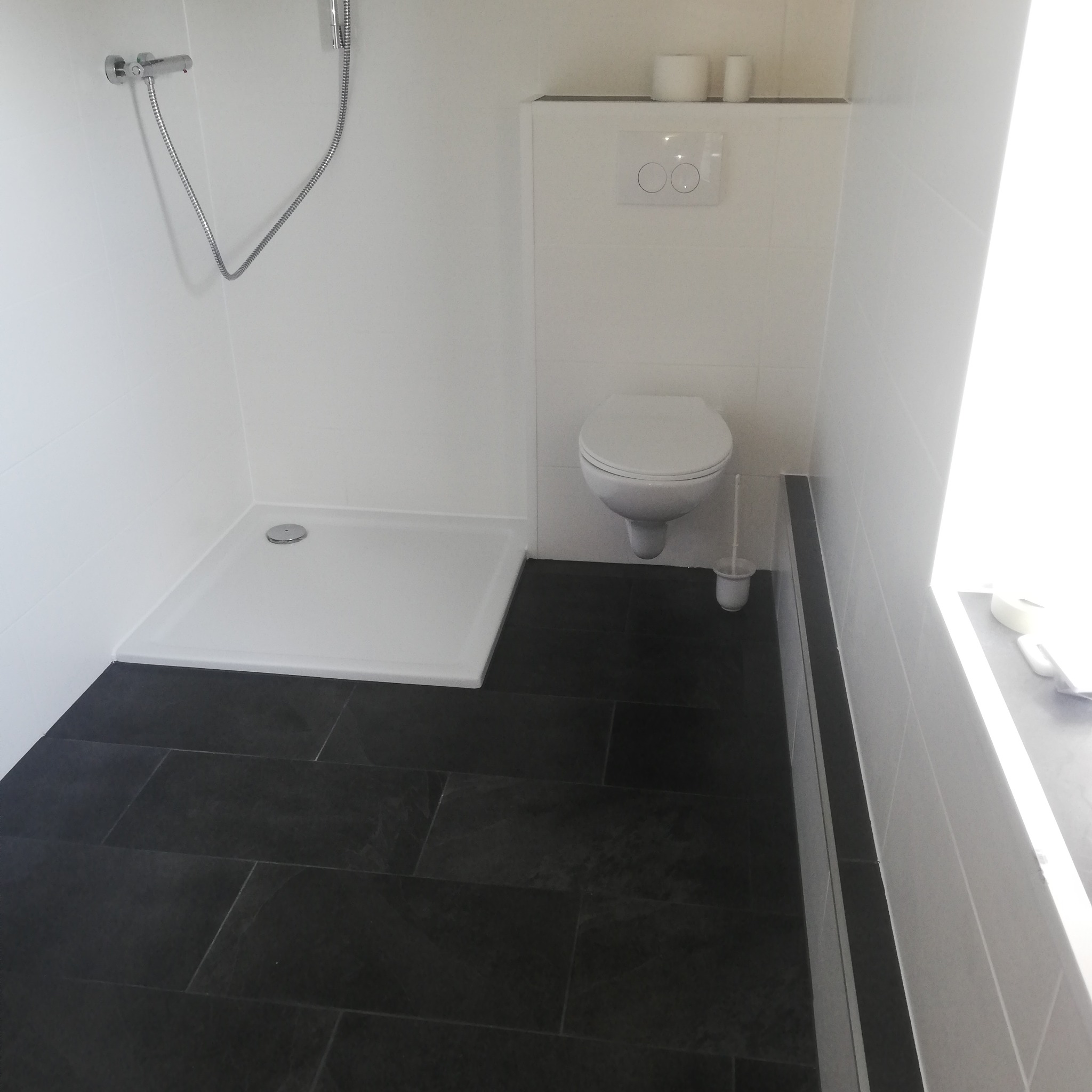 Nowoczesna łazienka po remoncie: białe płytki ścienne, czarna podłoga z płytek imitujących kamień, kabina prysznicowa i podwieszana toaleta.