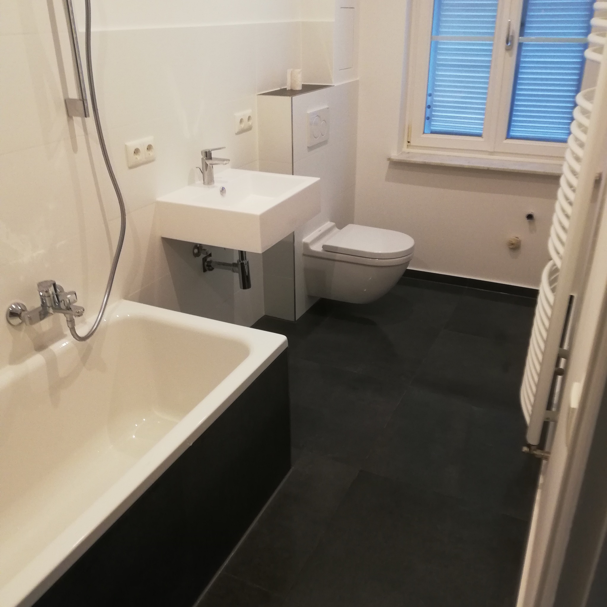 Minimalistyczna łazienka po remoncie: biała armatura (wanna, umywalka, toaleta), czarna podłoga, okno z żaluzjami. Widok na baterię wannową i grzejnik.