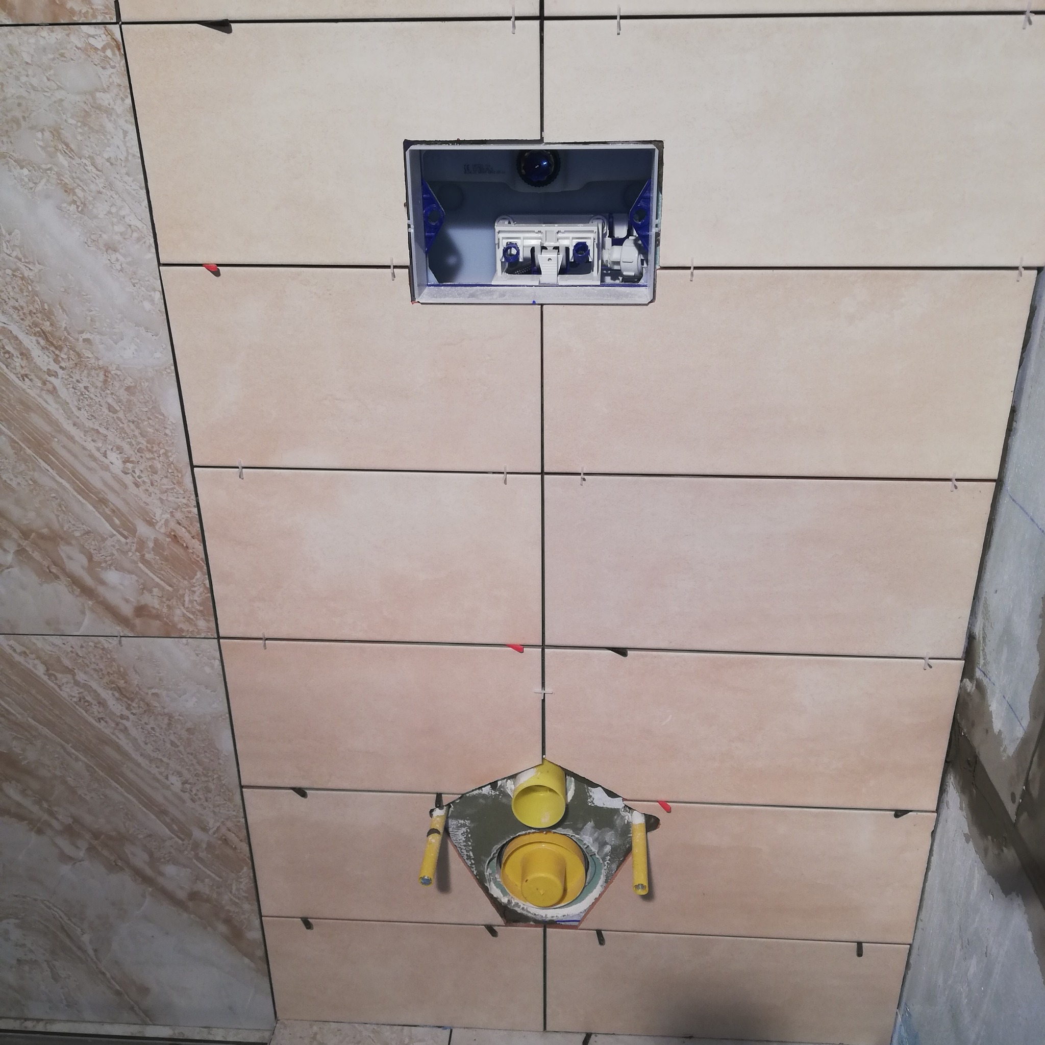 Układanie beżowych płytek w łazience, widoczne elementy instalacji podtynkowych WC i bidetu, krzyżyki dystansowe. Fragment ściany z marmurową okładziną po lewej.