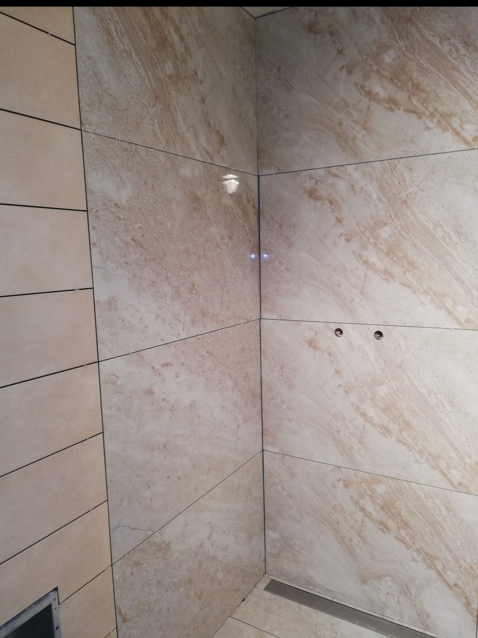 Beżowe płytki imitujące marmur, ułożone w rogu kabiny prysznicowej z odpływem liniowym. Widoczne otwory na baterię i krzyżyki dystansowe.