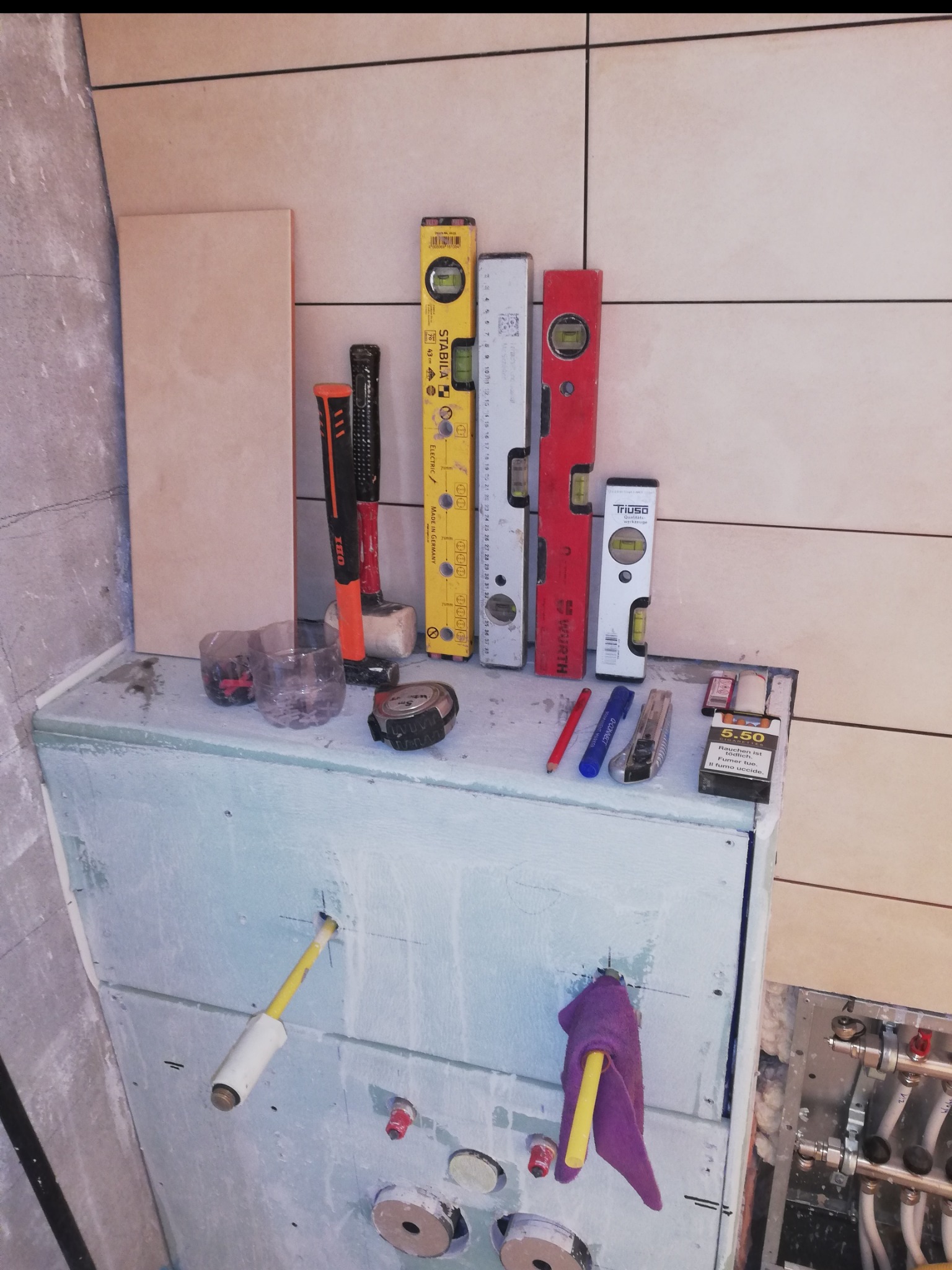 Przygotowanie do układania płytek w łazience: poziomice, narzędzia glazurnicze i fragment instalacji wodnej widoczne na tle częściowo wykończonej ściany.