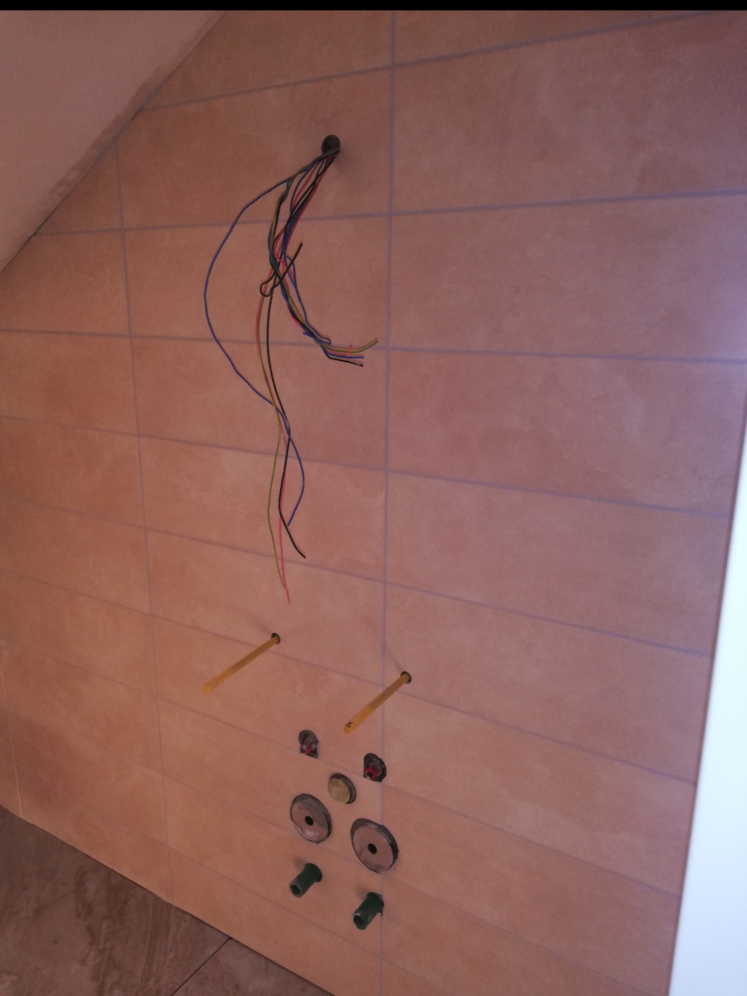 Ściana w łazience z beżowymi płytkami, wystającymi przewodami elektrycznymi i elementami instalacji wodnej przed montażem armatury. Widoczne pionowe i poziome fugi.