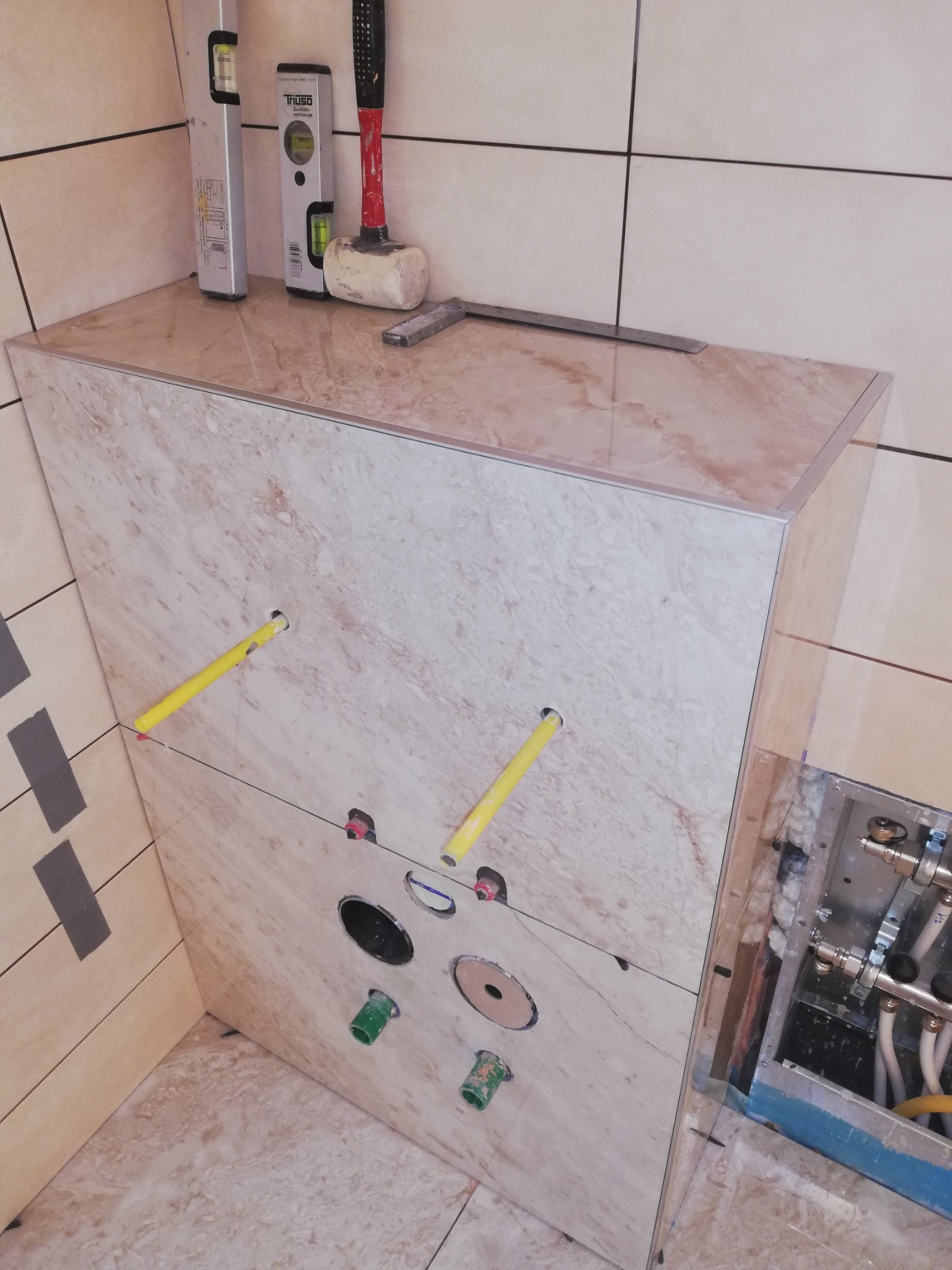 Obudowa stelaża WC z płytek imitujących marmur, z widocznymi przyłączami wodno-kanalizacyjnymi. Poziomica, młotek i szpachelka na górze. Detal wykończenia łazienki.