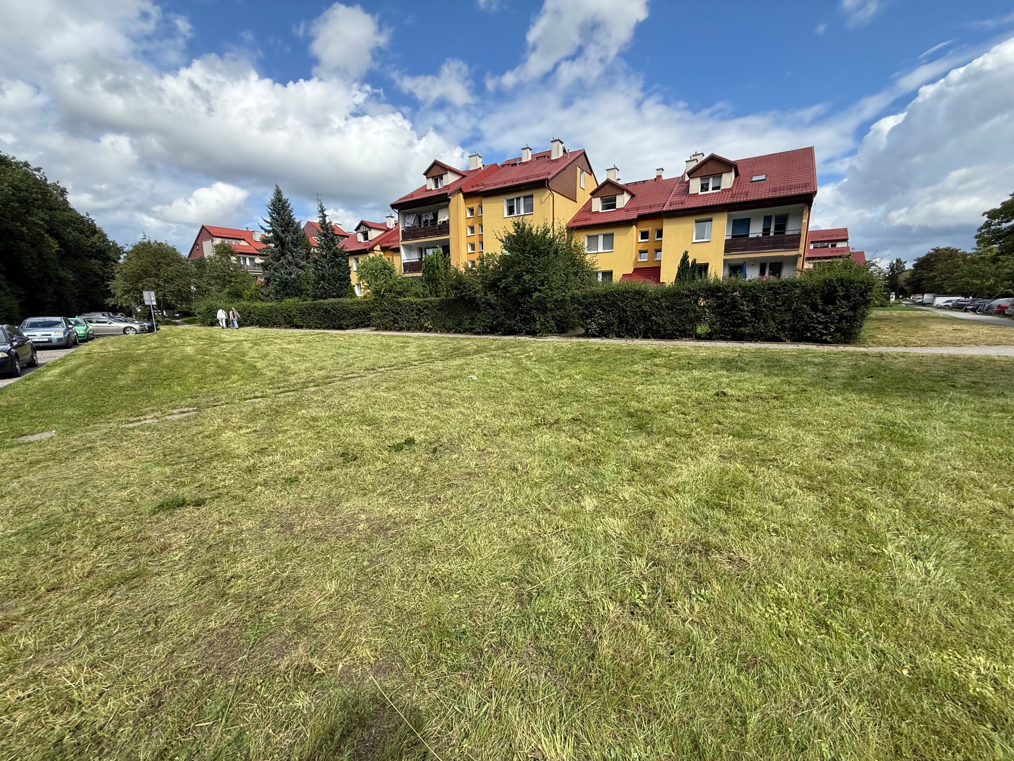 Żółty budynek mieszkalny z czerwonym dachem, otoczony zielenią trawnika i krzewów, pod błękitnym niebem z chmurami. Widoczny fragment świeżo skoszonej trawy.