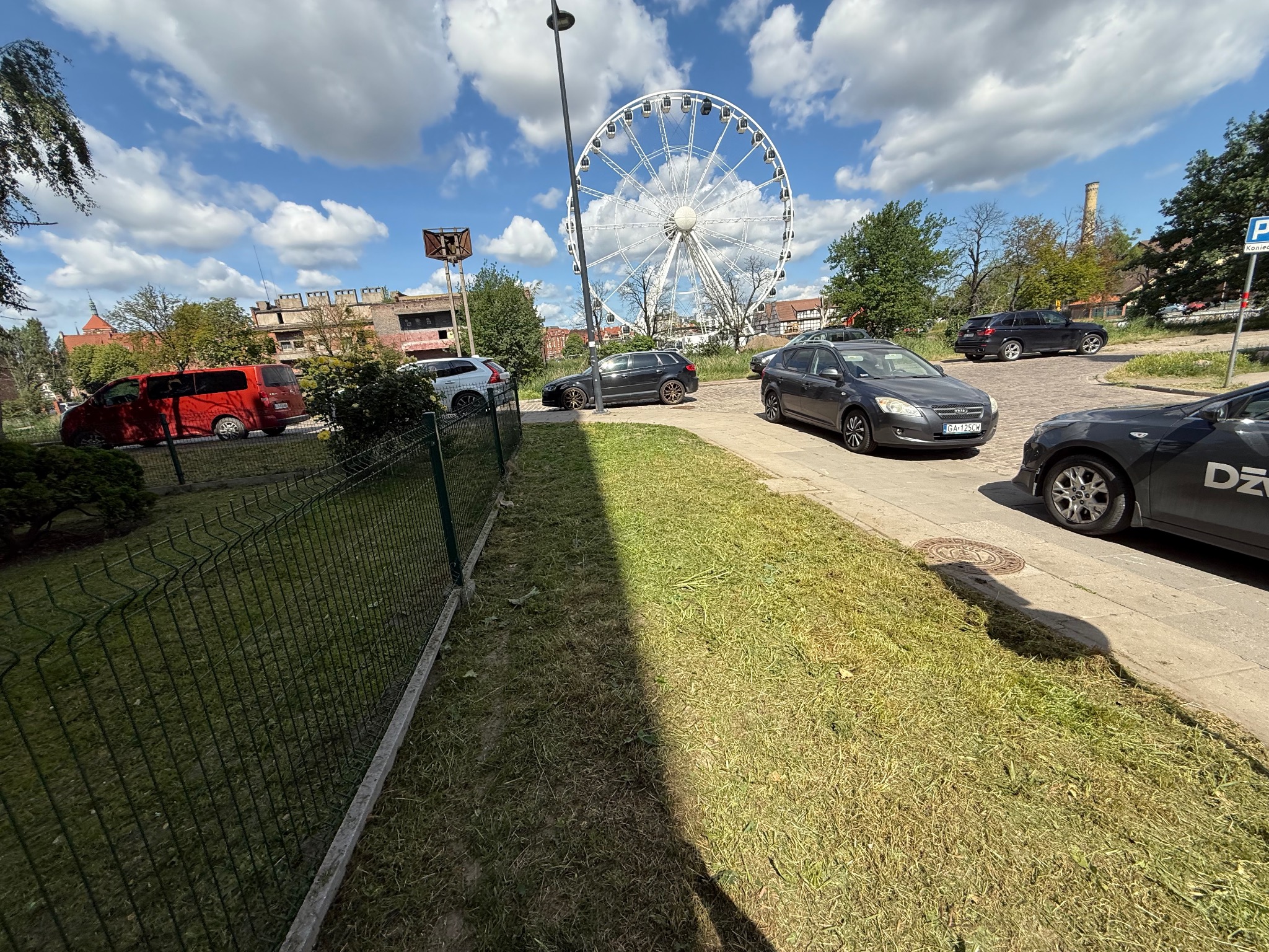 Skoszona trawa przy parkingu miejskim z diabelskim młynem w tle. Widoczne samochody i metalowe ogrodzenie.