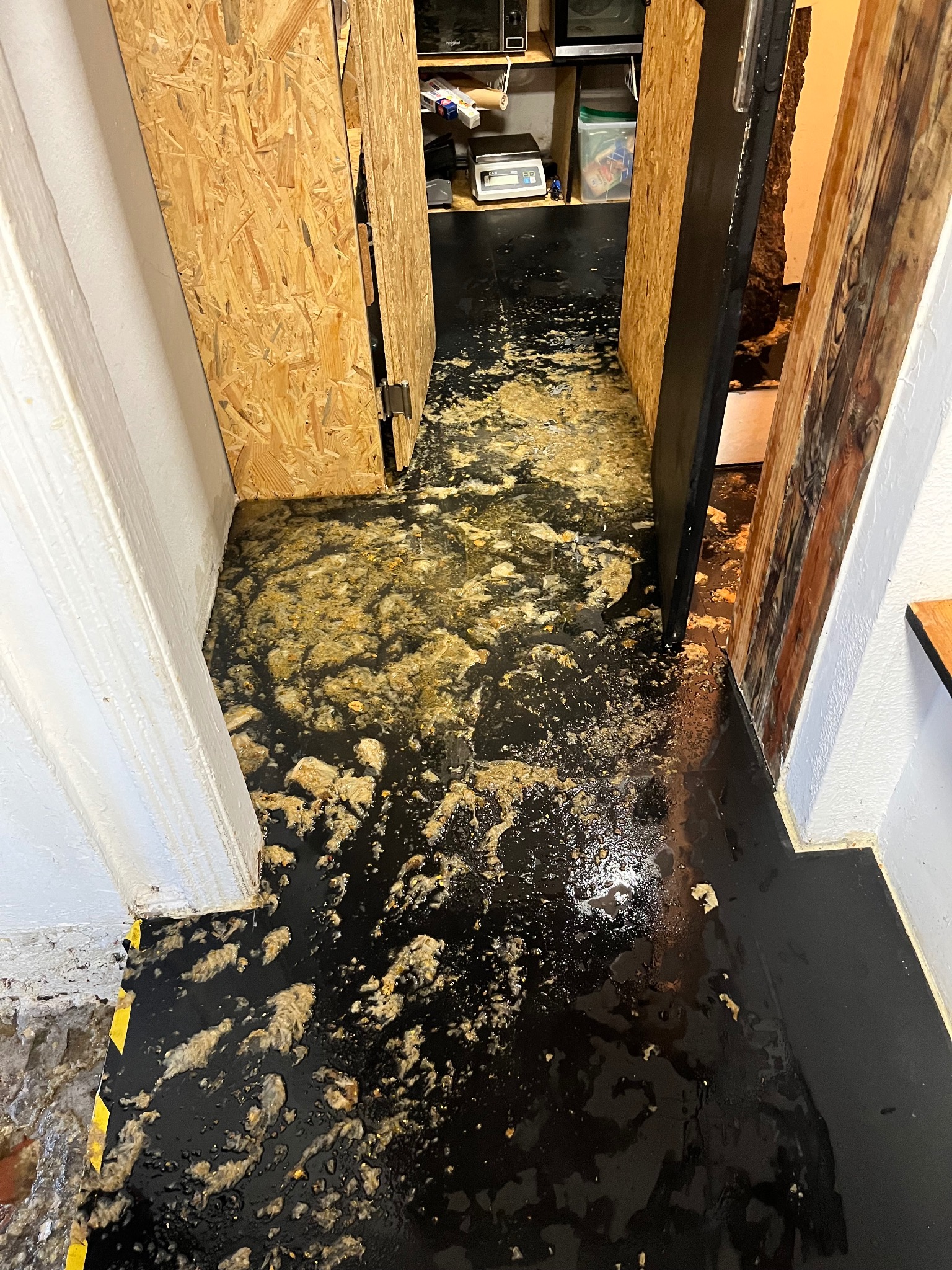 Zanieczyszczona, czarna podłoga w korytarzu z otwartymi drzwiami. Widoczne plamy i zabrudzenia. Fragment ściany z żółto-czarnym pasem na dole.