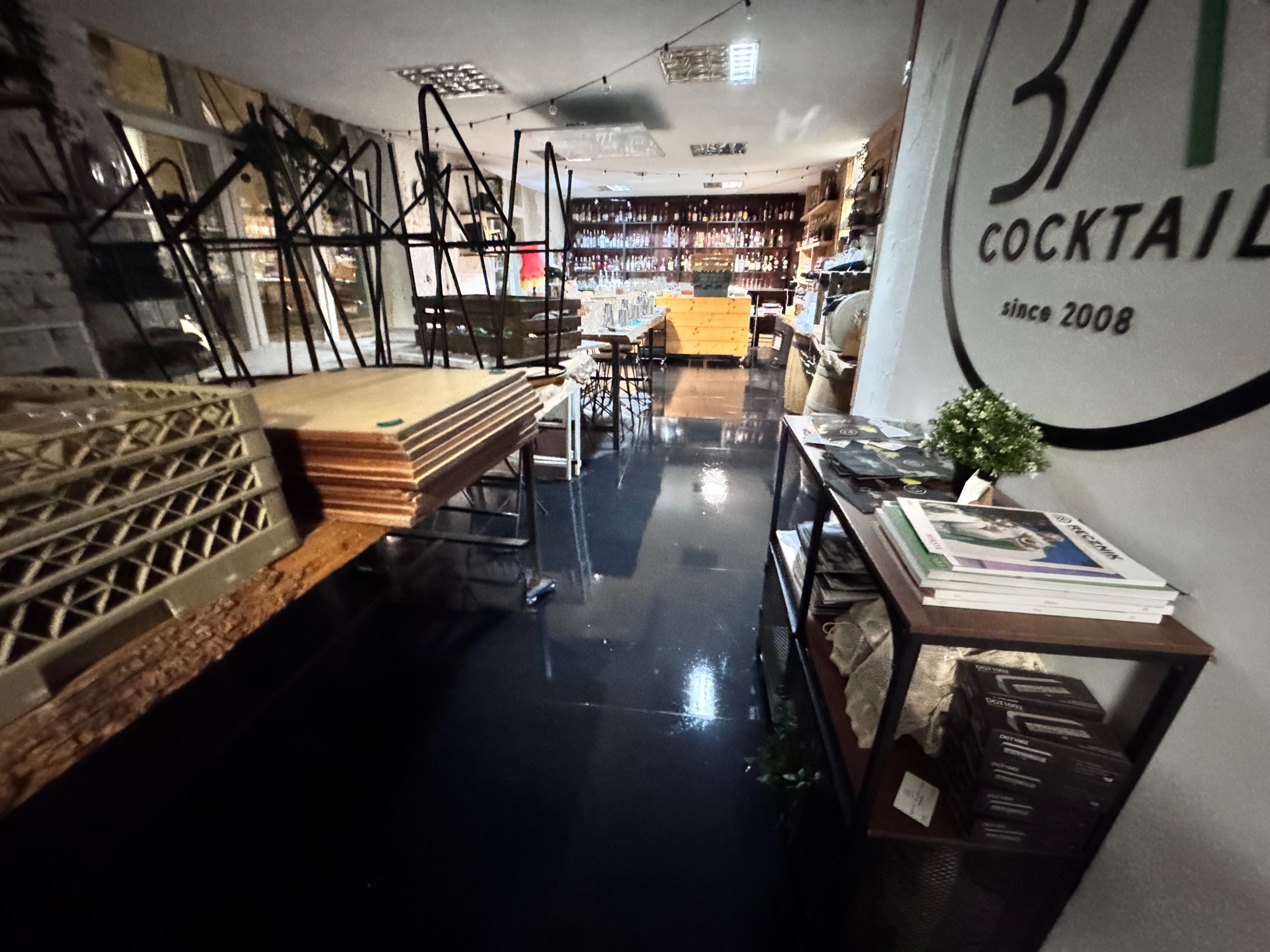 Wnętrze lokalu z barem, przygotowane do sprzątania. Widoczne krzesła na stołach, bar z butelkami w tle i logo '3/1 COCKTAIL' na ścianie.
