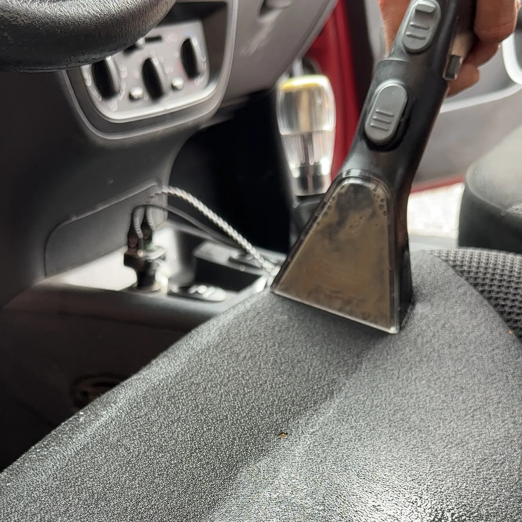 Czyszczenie tapicerki samochodowej parownicą. Dysza urządzenia przesuwa się po szarym fotelu, w tle deska rozdzielcza i dźwignia zmiany biegów.