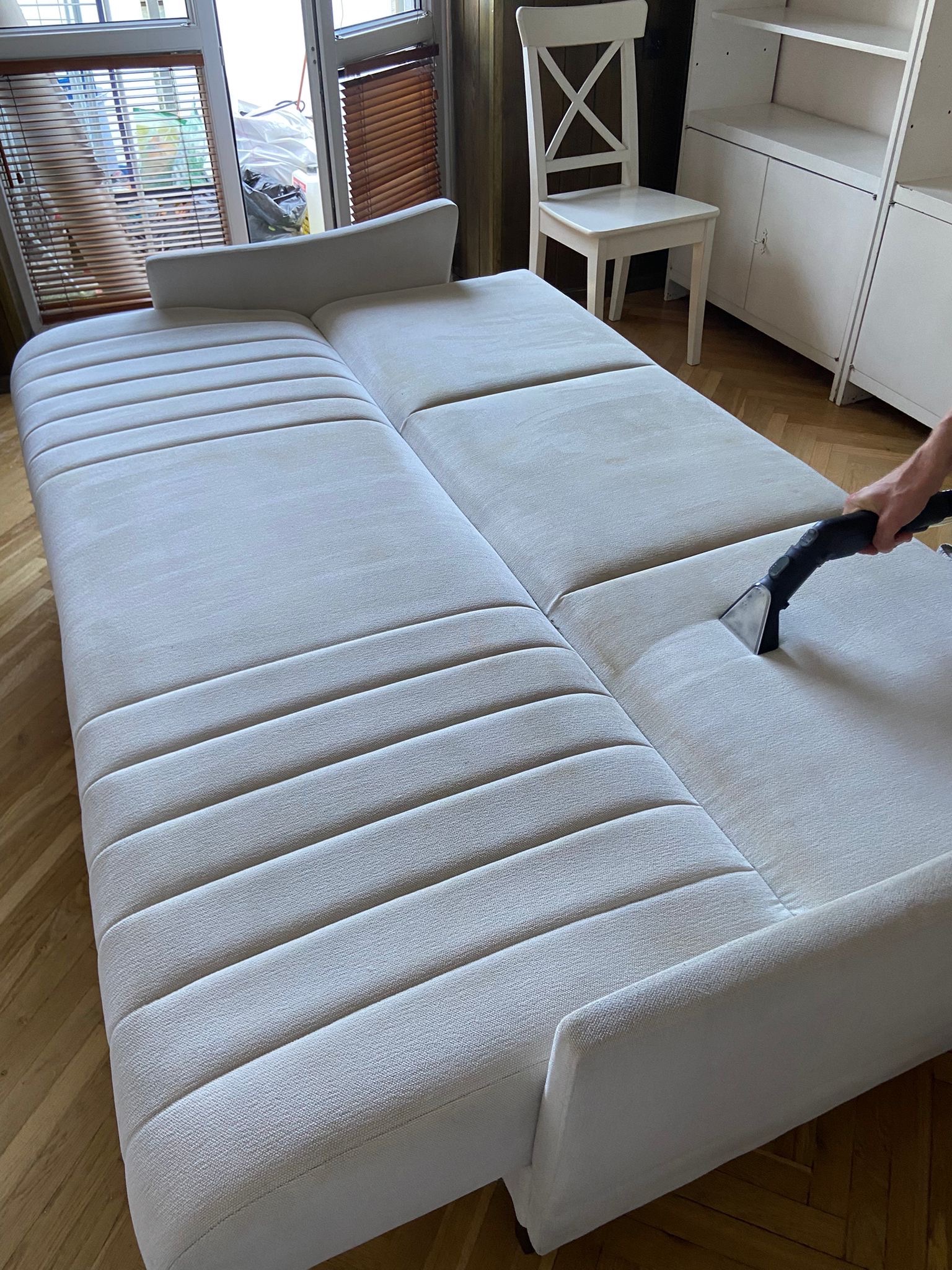 Pranie jasnej, rozkładanej sofy z wyraźnymi przeszyciami, za pomocą specjalistycznego odkurzacza. Widoczny fragment parkietu i białe meble w tle.