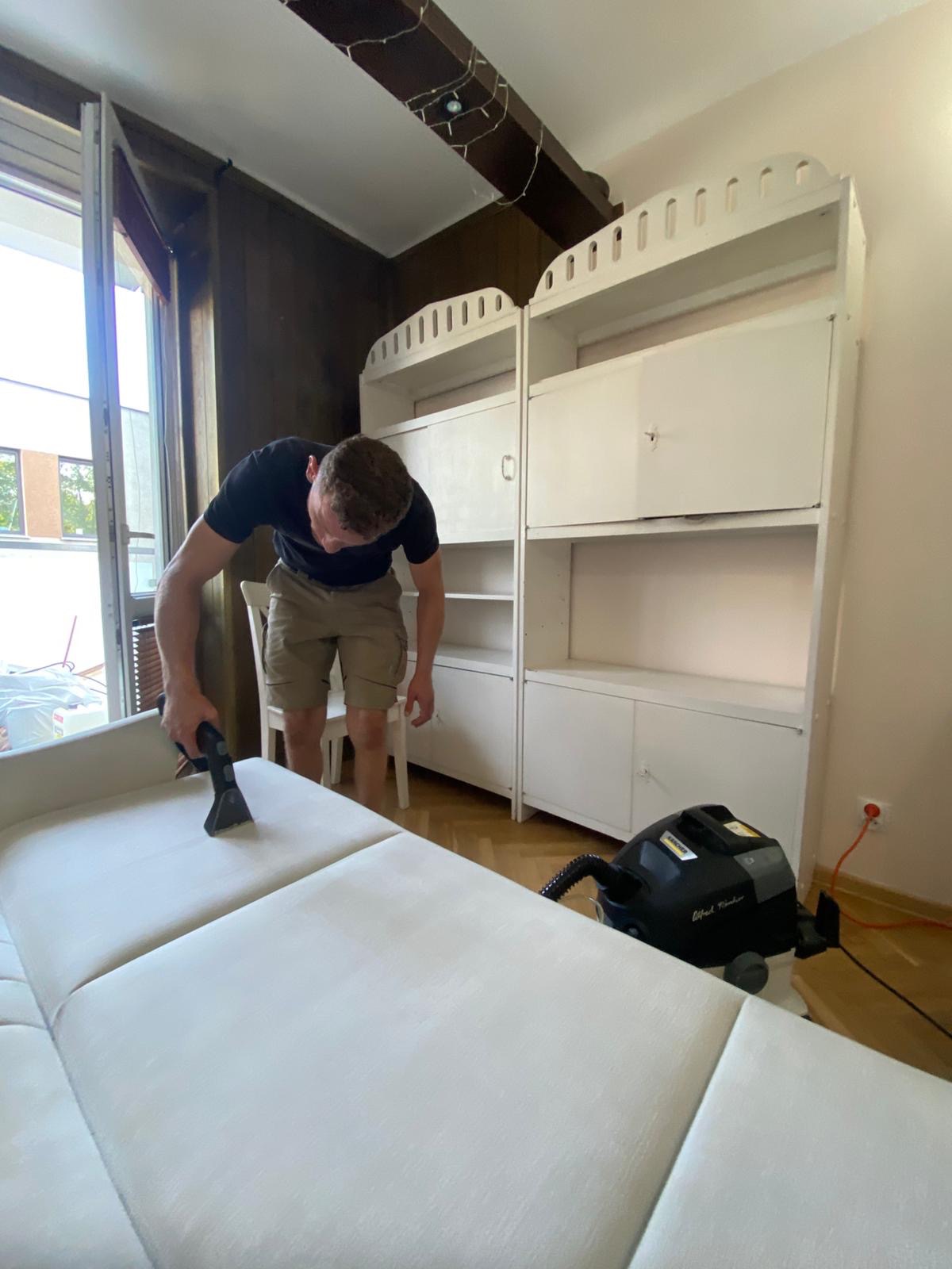 Pranie jasnej kanapy metodą ekstrakcyjną. Mężczyzna czyści tapicerkę za pomocą odkurzacza piorącego, obok białe regały i otwarte okno.