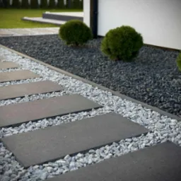 Nowoczesna ścieżka z ciemnych płyt na jasnym żwirze, obok rabata z ciemnym grysem i kulistymi krzewami. Minimalistyczny design ogrodu.