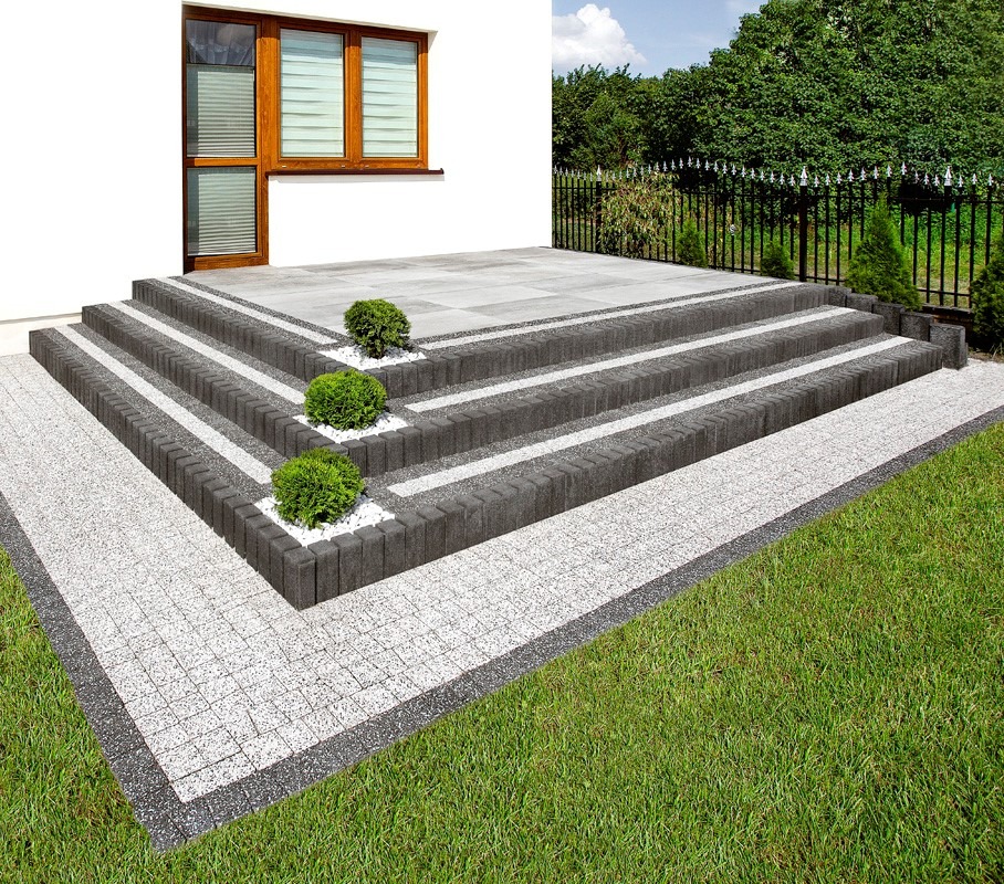 Schody z kostki brukowej prowadzące do domu, z małymi krzewami ozdobnymi i jasną kostką na podjeździe, otoczone zielonym trawnikiem. Eleganckie wykończenie.