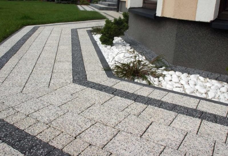 Kremowa kostka brukowa z ciemnymi pasami, tworząca ścieżkę przy domu. Obok rabata z białymi kamieniami i roślinami. Estetyczne wykończenie terenu.