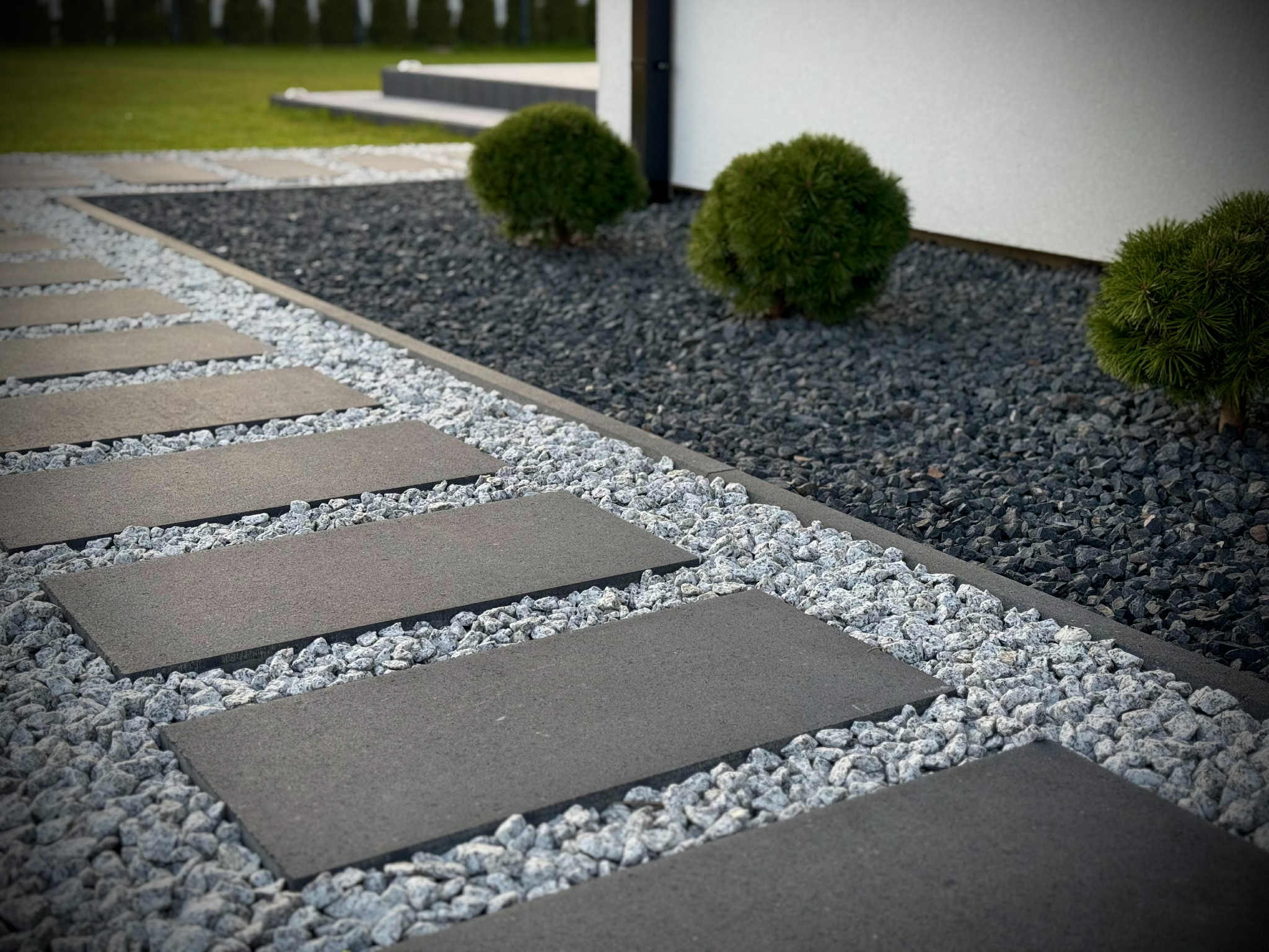 Nowoczesna ścieżka z ciemnych płyt na jasnym żwirze, obok rabata z ciemnym grysem i kulistymi krzewami. Minimalistyczny design ogrodu.