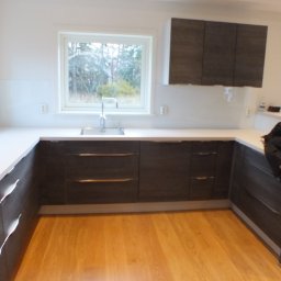 BRACIA PO FACHU SPÓŁKA Z OGRANICZONĄ ODPOWIEDZIALNOŚCIĄ - Nowoczesna kuchnia z drewnianą podłogą, szarymi szafkami z poziomymi uchwytami i białym blatem. Okno z widokiem na zieleń. Minimalistyczny design.