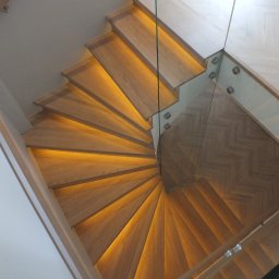 BRACIA PO FACHU SPÓŁKA Z OGRANICZONĄ ODPOWIEDZIALNOŚCIĄ - Drewniane schody spiralne z podświetleniem LED, widok z góry. Szklana balustrada z metalowymi mocowaniami. Parkiet ułożony w jodełkę. Nowoczesne wykończenie wnętrza.
