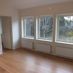 BRACIA PO FACHU SPÓŁKA Z OGRANICZONĄ ODPOWIEDZIALNOŚCIĄ - Jasne, puste wnętrze z drewnianą podłogą i trzema oknami z widokiem na drzewa. Białe ściany, grzejniki pod oknami, otwarte drzwi do sąsiedniego pomieszczenia.
