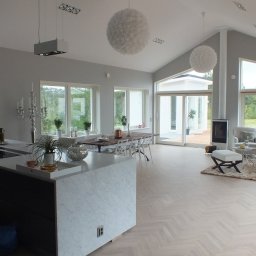 BRACIA PO FACHU SPÓŁKA Z OGRANICZONĄ ODPOWIEDZIALNOŚCIĄ - Jasne, przestronne wnętrze domu z parkietem w jodełkę, wyspą kuchenną z marmurowym wykończeniem i widokiem na basen przez duże okna.