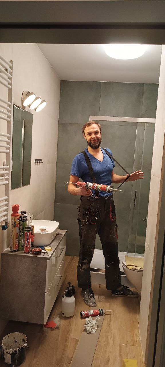 Remont łazienki: mężczyzna z pistoletem do silikonu na tle wykończonej łazienki z kabiną prysznicową, umywalką i grzejnikiem drabinkowym. Widoczne narzędzia i materiały.