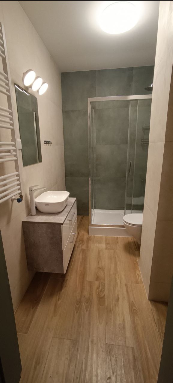 Nowoczesna łazienka po remoncie: kabina prysznicowa ze szklanymi drzwiami, umywalka nablatowa na szafce z szufladami, drewnopodobna podłoga i szare płytki.