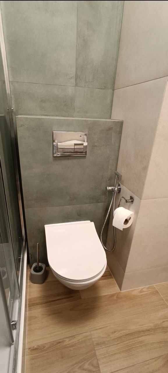 Nowoczesna toaleta w łazience z szarymi płytkami, podwieszaną miską WC i drewnopodobną podłogą. Widoczny chromowany przycisk spłuczki i bidetka.