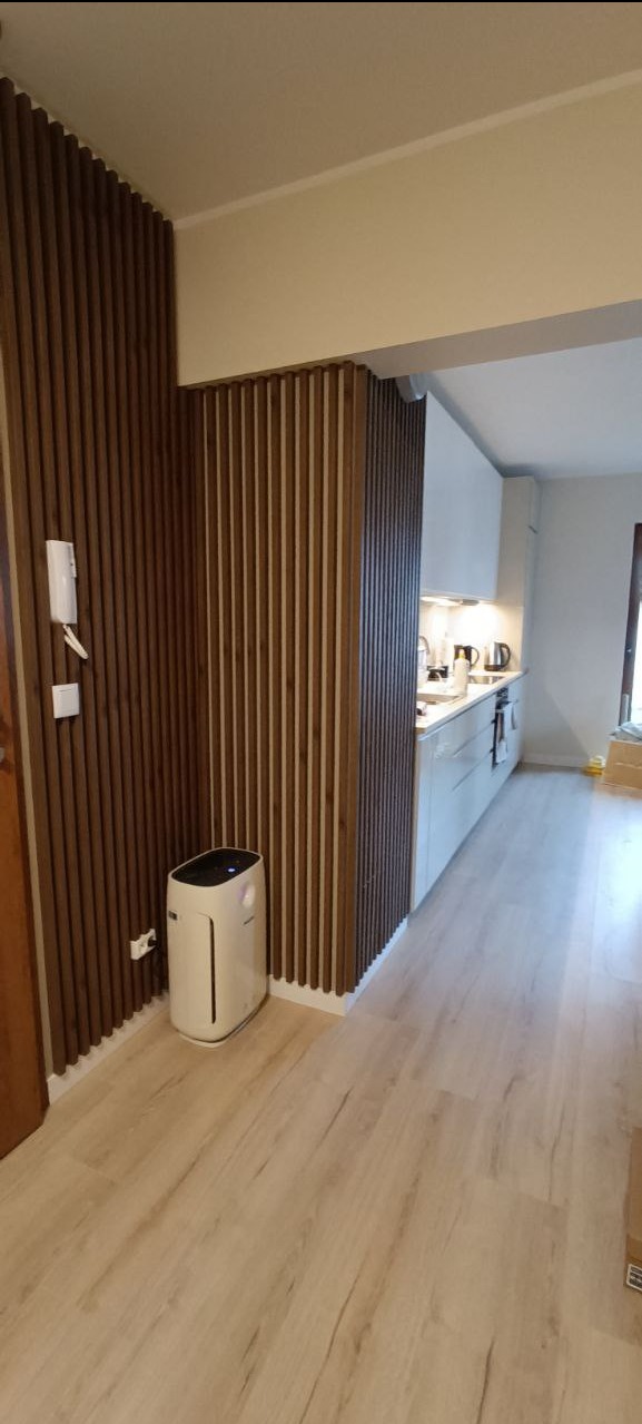 Nowoczesne wykończenie wnętrza: drewniane lamele na ścianie, jasna podłoga i widok na białą kuchnię. Minimalistyczny design z elementami dekoracyjnymi.