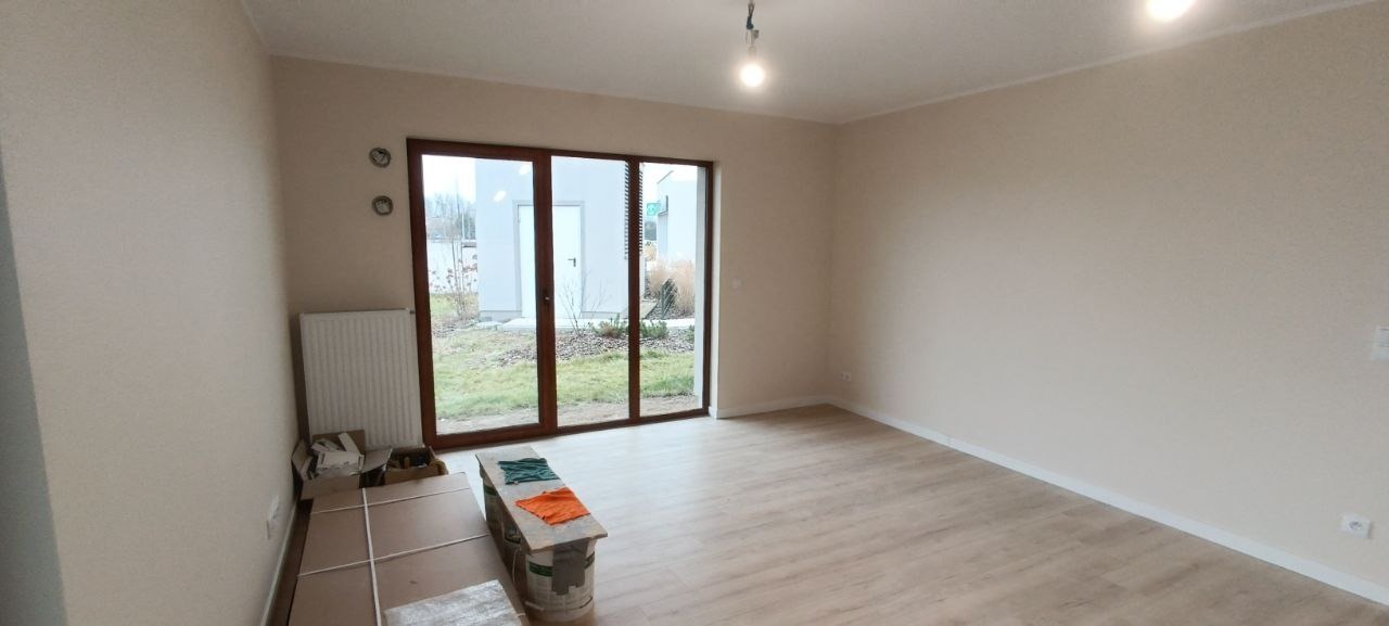 Jasne, pomalowane na beżowo wnętrze z dużym oknem balkonowym. Na podłodze panele, przy ścianie kartony i wiadro z farbą. Widoczne gniazdka elektryczne.