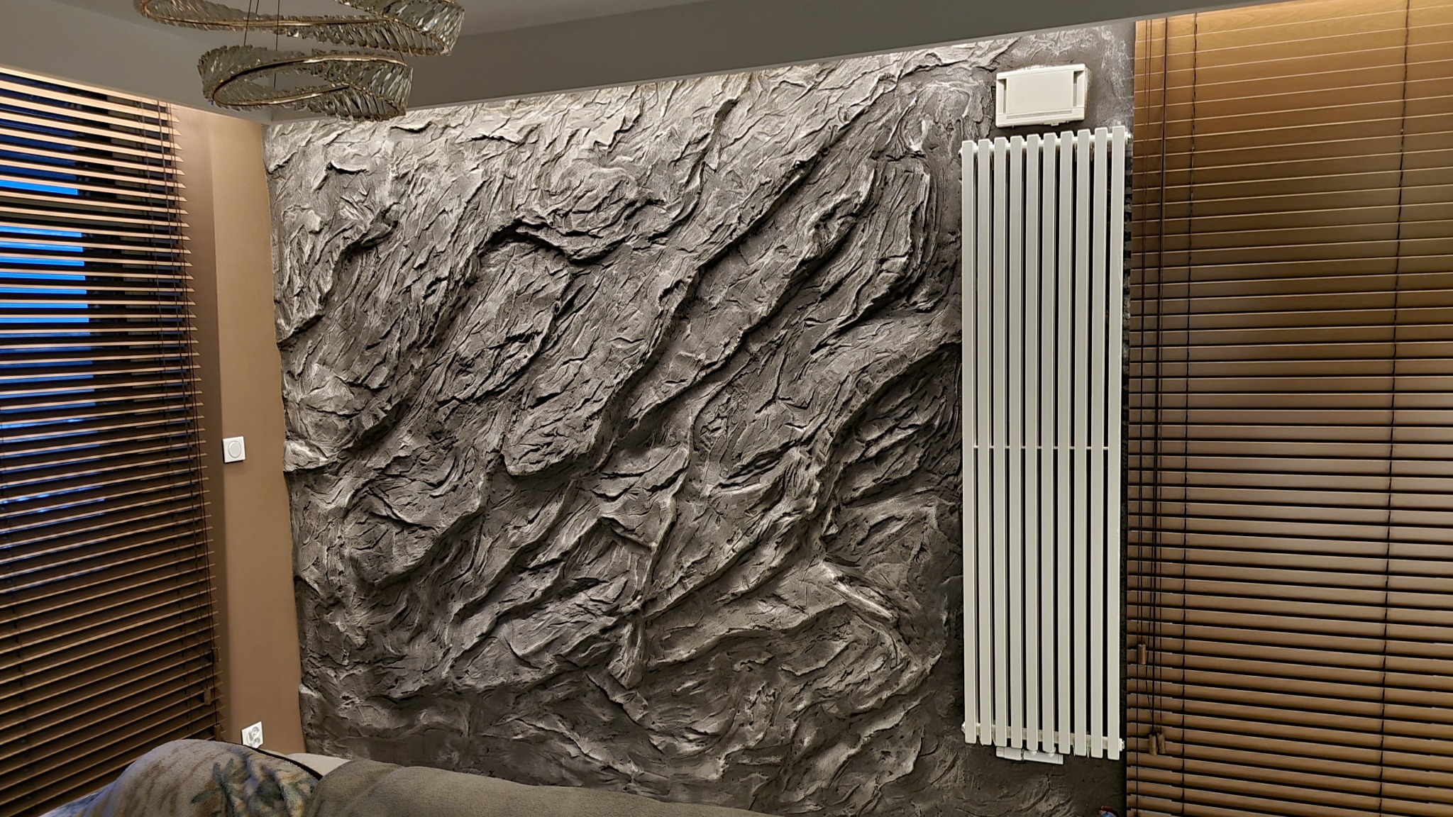 Dekoracyjna ściana o fakturze kamienia w szarościach, z białym grzejnikiem i drewnianymi żaluzjami. Nowoczesne oświetlenie sufitowe.