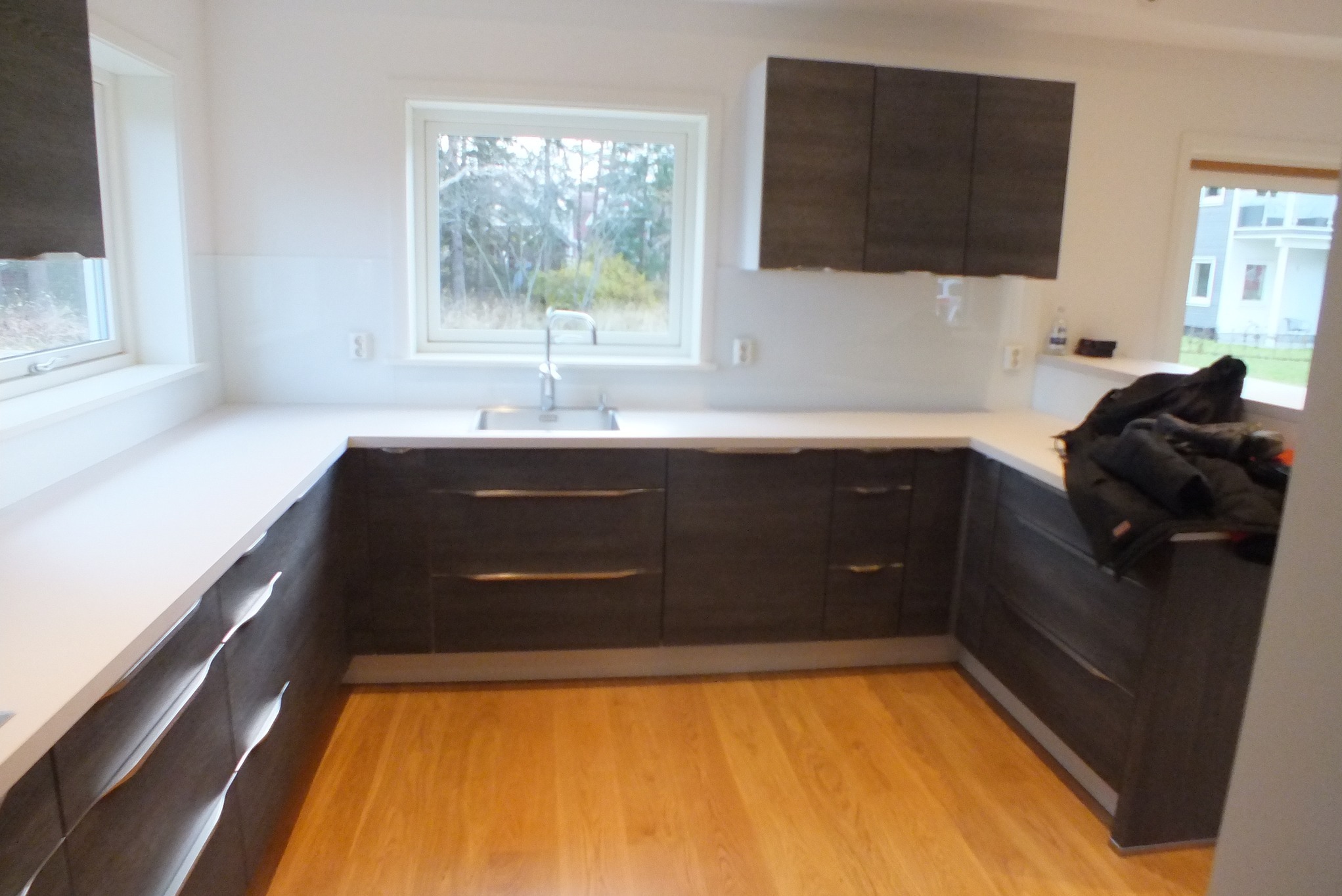 Nowoczesna kuchnia z drewnianą podłogą, szarymi szafkami z poziomymi uchwytami i białym blatem. Okno z widokiem na zieleń. Minimalistyczny design.