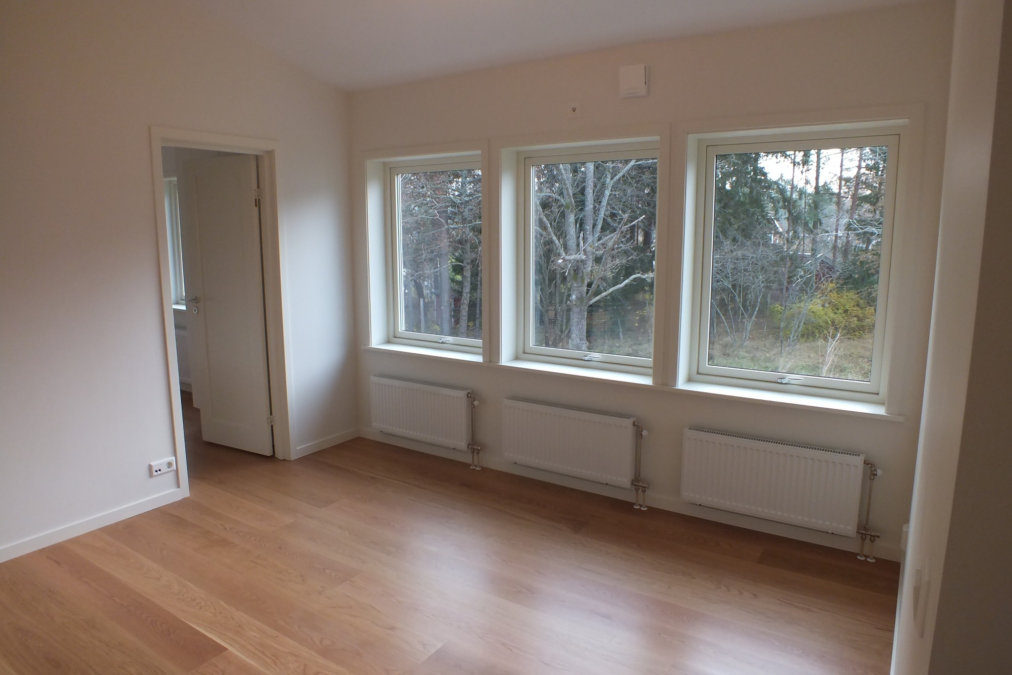 Jasne, puste wnętrze z drewnianą podłogą i trzema oknami z widokiem na drzewa. Białe ściany, grzejniki pod oknami, otwarte drzwi do sąsiedniego pomieszczenia.