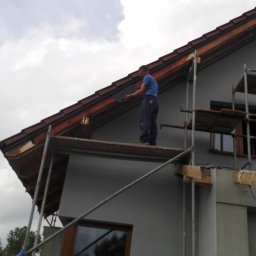 Robert Jaworski - Pracownik na rusztowaniu montuje elementy wykończeniowe pod okapem dachu domu. Widoczne drewniane elementy i szara elewacja budynku w trakcie prac.