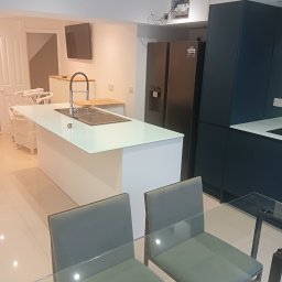 Robert Jaworski - Nowoczesna kuchnia w Małym Miłobądzu z wyspą i ciemnymi szafkami. Widoczne szklane krzesła i stół. Minimalistyczny design, jasne płytki podłogowe.