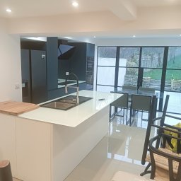 Robert Jaworski - Nowoczesna kuchnia z wyspą i widokiem na ogród przez duże okna. Minimalistyczny design, białe fronty, ciemne szafki i sprzęt AGD. Jasne płytki podłogowe.