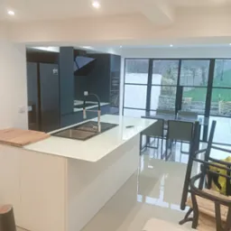 Nowoczesna kuchnia z wyspą i widokiem na ogród przez duże okna. Minimalistyczny design, białe fronty, ciemne szafki i sprzęt AGD. Jasne płytki podłogowe.