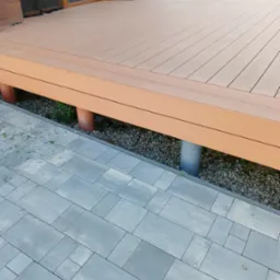 Wykończony taras z kompozytowych desek, wsparty na rurach, widok z boku na szarą kostkę brukową i rabatę z kamykami. Widoczne narzędzia do sprzątania.