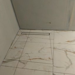 Robert Jaworski - Ułożone płytki imitujące marmur z odpływem liniowym w rogu łazienki. Widok z góry, neutralne oświetlenie.