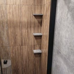 Robert Jaworski - Nowoczesna łazienka z drewnianą ścianą z pionowych paneli i trzema półkami z szarego betonu. Minimalistyczny design, ciepłe oświetlenie.