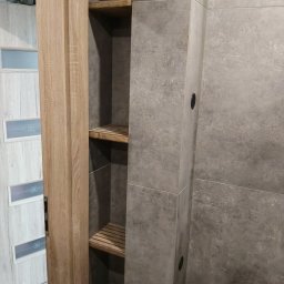 Robert Jaworski - Wykończona wnęka z drewnianymi półkami w szarej, nowoczesnej łazience z płytkami imitującymi beton. Widoczny fragment drzwi z drewnopodobną okleiną.