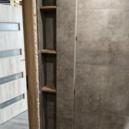 Robert Jaworski - Wykończona łazienka z szarą, matową glazurą i wnęką z drewnianymi półkami. Widoczny fragment ościeżnicy z pianką montażową i drzwi z przeszkleniem.