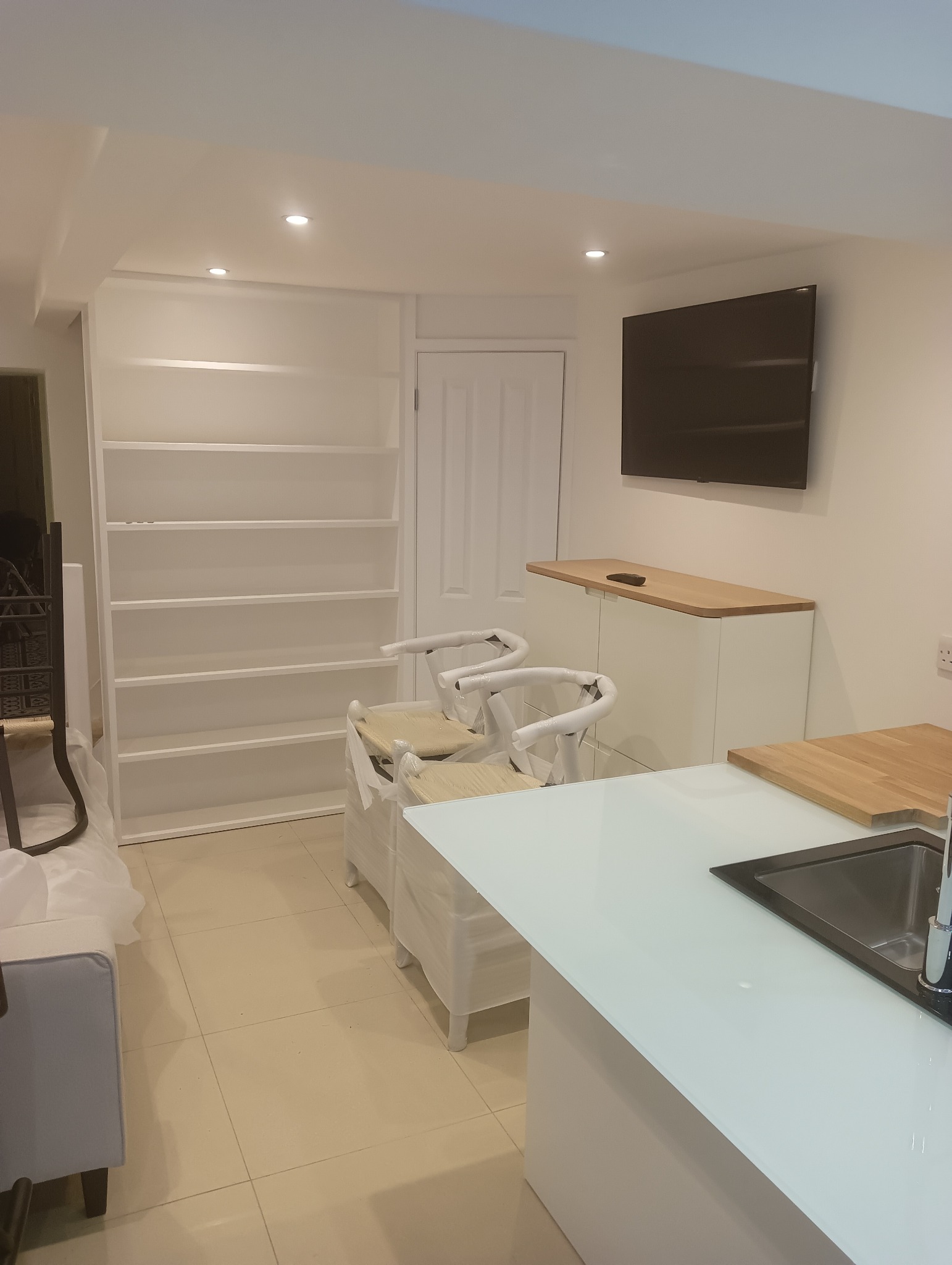 Jasna kuchnia po remoncie: białe meble, wyspa z blatem, krzesła w folii ochronnej i półki w zabudowie, minimalistyczny design, świeżo po wykończeniu.