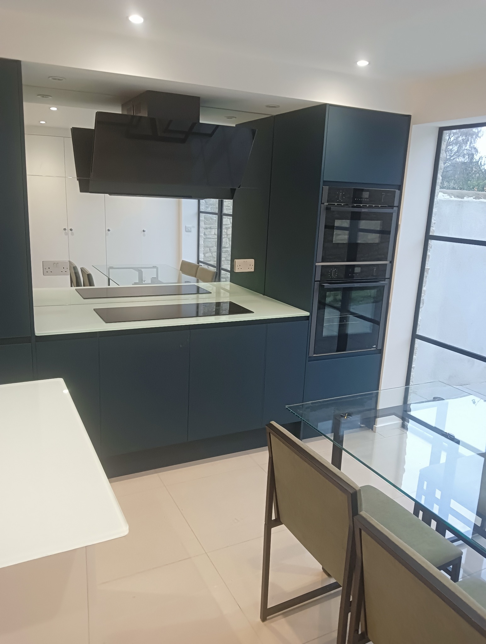 Nowoczesna, minimalistyczna kuchnia w odcieniach zieleni i bieli. Zabudowa kuchenna z piekarnikiem i płytą indukcyjną. Szklany stół z krzesłami w tle.