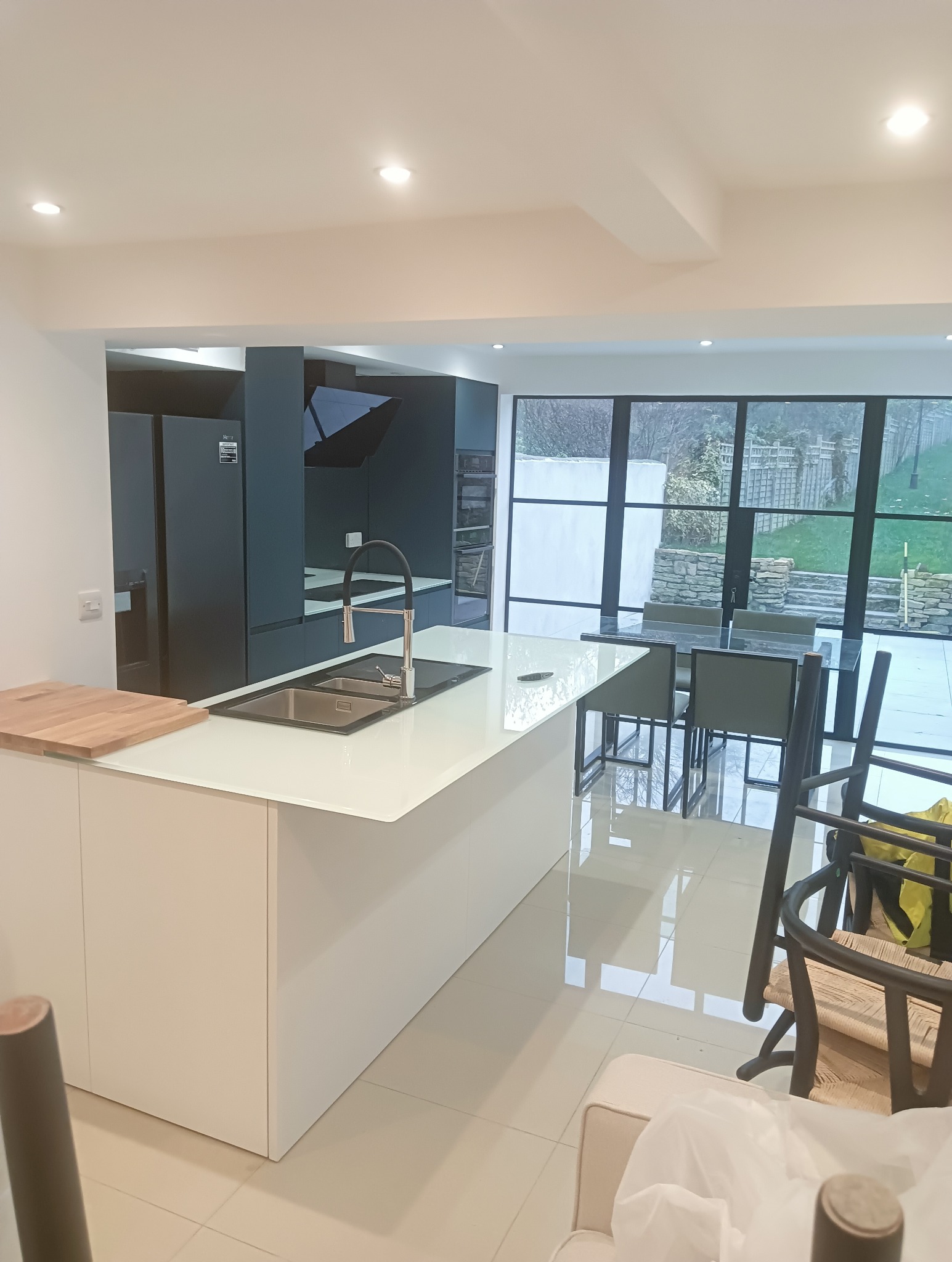 Nowoczesna kuchnia z wyspą i widokiem na ogród przez duże okna. Minimalistyczny design, białe fronty, ciemne szafki i sprzęt AGD. Jasne płytki podłogowe.