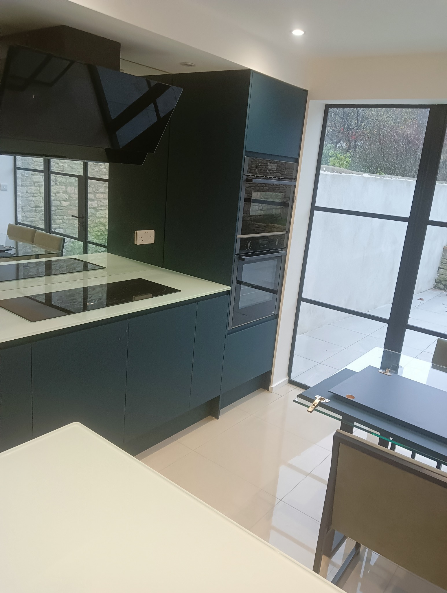 Nowoczesna, ciemnozielona zabudowa kuchenna z białym blatem i wbudowanymi sprzętami AGD. Duże okno z czarnymi szprosami doświetla wnętrze.