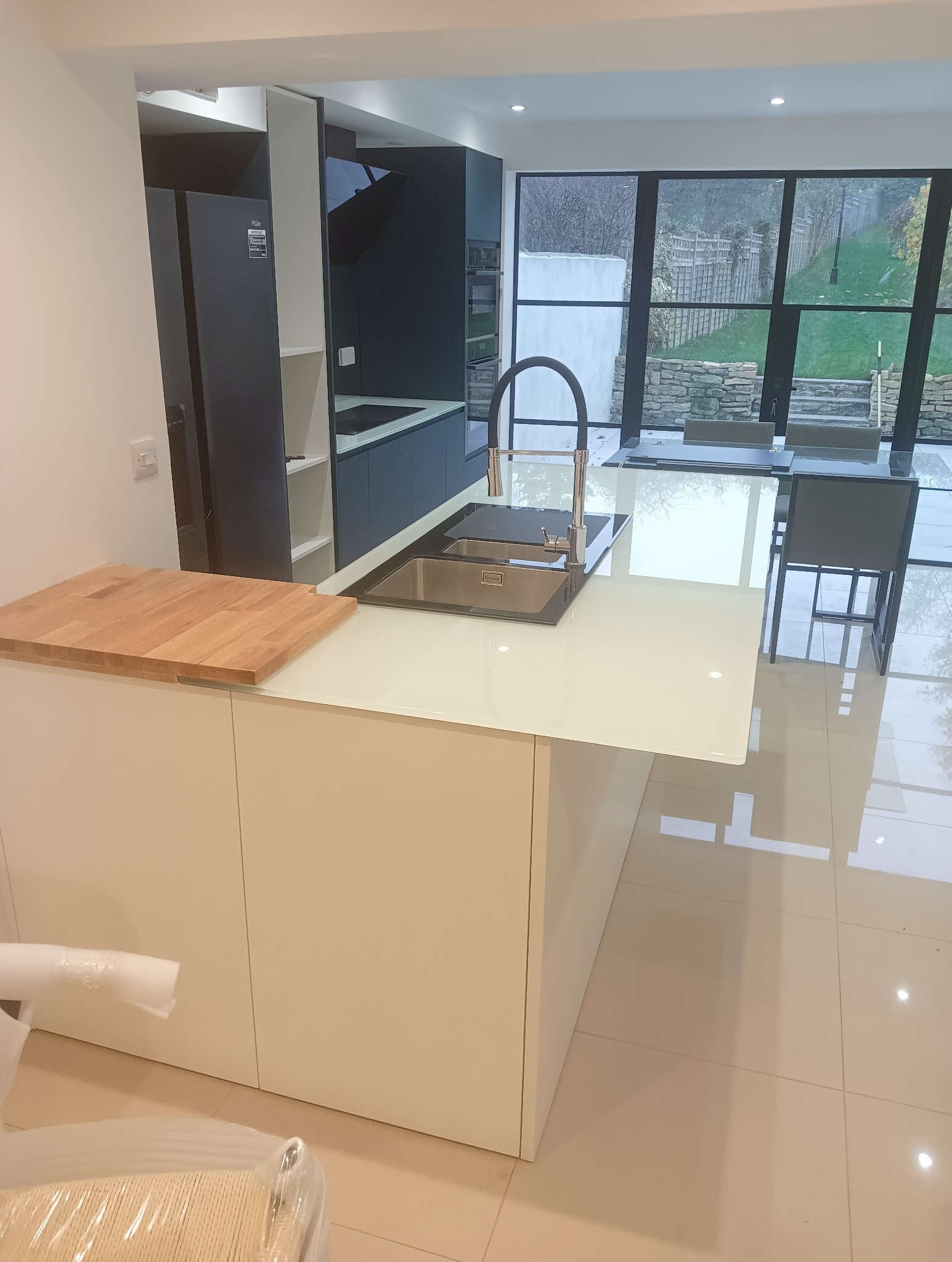 Nowoczesna kuchnia z wyspą i białym blatem, czarne szafki w tle, okno z widokiem na ogród. Uwagę przyciąga drewniany element na wyspie.